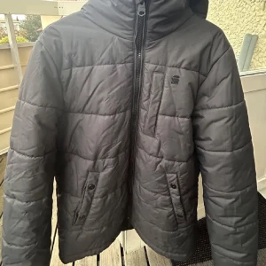 Svart pufferjacka från G-Star RAW L - Säljer en svart pufferjacka från G-Star RAW i storlek L. Jackan har huva, dragkedja framtill, två fickor med knappar och en bröstficka. Snygg quiltad design och perfekt för kyliga dagar. Klassisk logga på bröstet och stilren look.