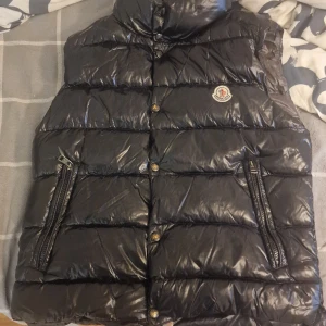 Svart dunväst från Moncler - Snygg svart dunväst från Moncler med glansig finish och quiltad design. Västen har två fickor med dragkedja, hög krage och klassisk Moncler-logga på bröstet. Insidan har en unik serietidningsetikett. Perfekt för att addera lite lyx till din look.