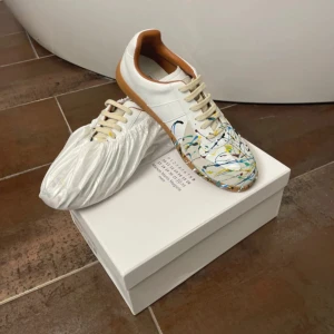 Maison Margielas sneakers med färgstänk - Margielas sneakers i vitt skinn med beige detaljer och gummisula. Ovandelen har handmålade färgstänk i blått, gult, grönt och rött för en unik look. Klassisk rund tå, snörning och låg profil. Insidan är fodrad i brunt skinn för extra komfort.