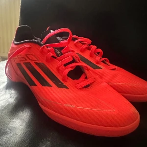 Röda Adidas fotbollsskor med snörning - Säljer ett par snygga inomhus skor från Adidas i starkt röd färg med svarta detaljer och klassiska tre ränder på sidan. Skorna har snörning och är tillverkade i syntetmaterial med mesh för bra ventilation. Perfekt för dig som vill sticka ut på planen.