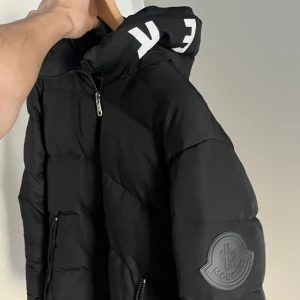 Svart Moncler pufferjacka med huva - Svart pufferjacka från Moncler med stor huva och vit logotyptext. Jackan har två sidofickor med dragkedja, quiltad design och Moncler-märke på ärmen. Helt ny oanvänd, kom med prisförslag:). 