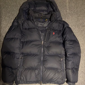 Svart pufferjacka från Polo Ralph Lauren - Snygg svart pufferjacka från Polo Ralph Lauren (knappt använd) med huva och klassisk röd logga på bröstet. Jackan har flera dragkedjeförsedda fickor och quiltad design som ger en cool och sportig vibe. Perfekt för kalla dagar och enkel att matcha med jeans eller hoodies.