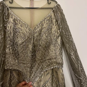 Glittrig långklänning med paljetter - Säljer en lyxig långklänning i champagnefärg med silverglittriga paljetter och mönster. Klänningen har v-ringning, långa transparenta ärmar och är täckt av detaljerade paljettbroderier. Perfekt för speciella tillfällen och maxad glamour.