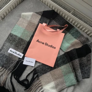 Rutig halsduk Acne Studios ull - Snygg och stor rutig halsduk från Acne Studios i mjuk ullmix. Färgerna är svart, grå, beige, vit och mintgrön. Klassisk design med fransar i kanterna och Acne Studios-logga. Perfekt att svepa runt halsen för en cool look.