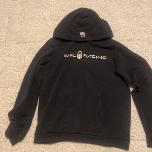  Sail Racing hoodie i bra kondition  - Svart hoodie från Sail Racing med stor vit logga på bröstet och en mindre logga på huvan. Tröjan har en klassisk känguruficka framtill och är tillverkad i mjuk bomull med fleece på insidan. Perfekt för dig som gillar sportig och clean stil.