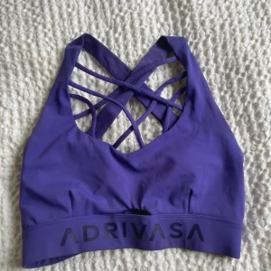 Lila sportig linnetopp från Adrivasa - Snygg lila tränings topp från Adrivasa med coola korsade band i ryggen och bred resår med logga framtill. Toppen är ärmlös och har en tight passform som passar perfekt till träning. Materialet är mjukt och stretchigt syntet för maximal komfort. Endast testad!