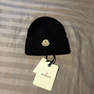 Svart Moncler mössa i ull - Svart ribbstickad mössa från Moncler med klassisk uppvikt kant och broderad logotyp framtill. Tillverkad i mjuk ull som håller dig varm under kyliga dagar. Perfekt accessoar för en stilren och exklusiv look.
