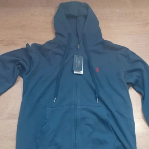 Marinblå hoodie från Polo Ralph Lauren - Snygg marinblå hoodie från Polo Ralph Lauren med dragkedja och huva. Tröjan har snörning i huvan, fickor framtill och det klassiska röda Polo-logot broderat på bröstet. Tillverkad i mjuk bomull för skön komfort. Custom fit-modell som sitter riktigt bra.