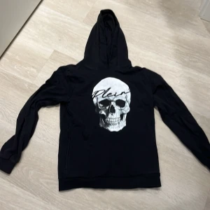 Philip Plein hoodie med tryck - Säljer en svart hoodie med ett stort vitt tryck på bröstet. Tröjan har huva och långa ärmar, samt ribbade muddar vid ärmslut och nederkant. Perfekt för dig som gillar streetwear och vill ha en cool look.