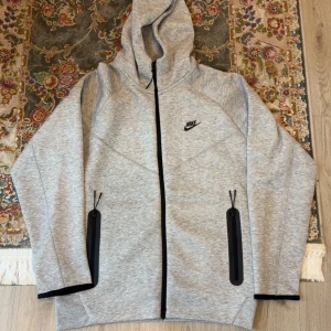 Grå zip-hoodie från Nike - Snygg grå zip-hoodie från Nike med svart logga på bröstet. Jackan har hel dragkedja, två fickor med svarta detaljer och huva. Tillverkad i mjuk bomullsblandning, perfekt för chill eller träning. Svarta kantband vid ärmslut ger en clean look.