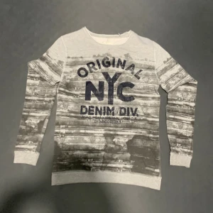 Grå NYC sweatshirt från Indigo Rebels - Snygg grå sweatshirt från Indigo Rebels med tryck 'ORIGINAL NYC DENIM DIV.' på bröstet. Tröjan har ett coolt, abstrakt mönster i svart och grått och är långärmad med ribbade muddar. Perfekt för dig som gillar streetstyle och vill sticka ut.
