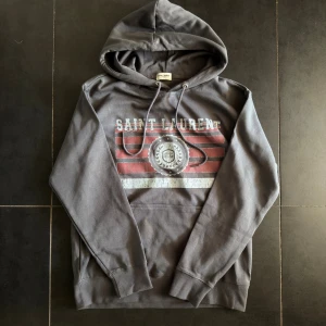 Grå Saint Laurent hoodie med tryck - Exklusiv Saint Laurent-hoodie i grått med logotryck och klassiska detaljer. Storlek M, passar som S. Sällsynt och stilren – en hoodie som verkligen sticker ut i skolan.