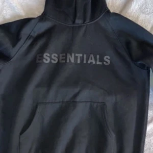 Svart Essentials hoodie  - Svart hoodie från Essentials med stor logga på bröstet och mindre tryck på ärmen. Klassisk känguruficka framtill och huva. Tröjan har en loose passform och är tillverkad i mjuk bomullsmix, perfekt för chill dagar.