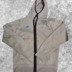 Grå Nike Air Max hoodie med dragkedja - Snygg grå hoodie från Nike Air Max med svart dragkedja och huva. Framtill finns blå Nike-logga och Air Max-tryck. Tröjan har långa ärmar och två fickor. Materialet känns mjukt och har en modern look, perfekt för dig som gillar sportig streetstyle.