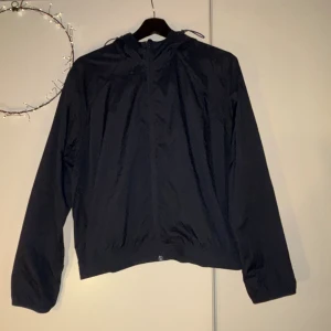 Mörkblå vindjacka från Filippa K Soft Sport - Mörkblå vindjacka från Filippa K Soft Sport i storlek S. Jackan har huva med snörning, hel dragkedja framtill och elastisk nederkant. Perfekt för dig som gillar stilren och sportig look. Materialet är lätt och syntetiskt, vilket gör den smidig att bära.