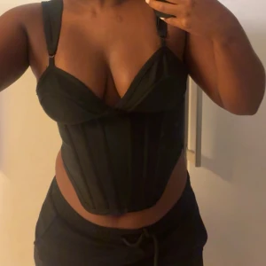 Svart korsett med dragkedja - Säljer en svart korsett med breda axelband och snygga kupor som ger en cool siluett. Toppen har markerade sömmar och dragkedja baktill för enkel på- och avtagning. Materialet är stadigt och ger en tight passform, perfekt för att lyfta din outfit.