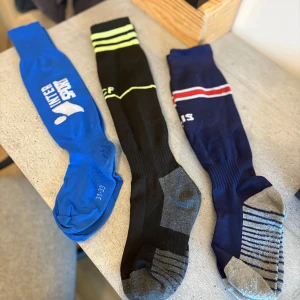 Tre par sportstrumpor, olika färger - Säljer tre par sportstrumpor i olika färger och stilar. Ett par är blå med vit Intersport-logga, ett par är svarta med neongula detaljer och grå förstärkning, och ett par är marinblå med röda och vita ränder samt grå-randig sula. Perfekta för träning eller fotboll.