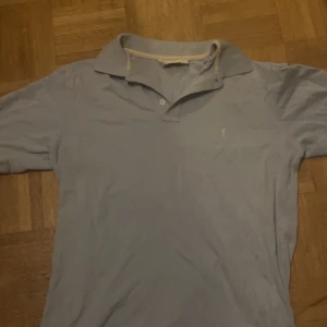 Ljusblå pikétröja från Yves Saint Laurent - Snygg ljusblå pikétröja från Yves Saint Laurent med klassisk krage och korta ärmar. Tröjan har diskret logga broderad på bröstet och knappar vid halsen. Perfekt för dig som gillar stilrena och exklusiva märken.