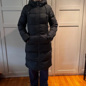 Svart lång dunjacka från Pajar Canada - Lång svart och tjock jacka från Pajar Canada med huva och dragkedja hela vägen. Jackan har quiltad design, två fickor med dragkedja och snygga detaljer vid ärmslut och midja. Perfekt för kalla dagar och riktigt varm. Klassisk look med diskret logga på ärmen. Figursydd. Jackan hade en päls som jag dessvärre tagit av och kastat då jag inte tyckte om den och hade inga planer på att sälja den sen men då den är i så fint skick och uppfattas av ögat som ny säljes den. Passar om man pendlar mellan S/XS