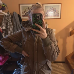 Beige pufferjacka med huva - Säljer en beige pufferjacka med huva och ribbade muddar. Jackan har dragkedja och knappar framtill samt praktiska fickor på sidorna. Perfekt för kyligare dagar och riktigt skön att bära.