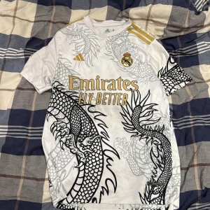 Real Madrid Vini Jr. draktröja Adidas - Säljer en fet Real Madrid fotbollströja med Vini Jr. och nummer 7 på ryggen. Tröjan är vit med guldiga detaljer och har ett sjukt snyggt drakmönster i svart och grått. Adidas-logga och klubbmärke på bröstet, korta ärmar och tillverkad i lätt polyester.