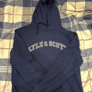 Marinblå hoodie från Lyle & Scott - Snygg marinblå hoodie från Lyle & Scott med stor logga tryckt över bröstet. Tröjan har huva med snörning och långa ärmar. Tillverkad i mjuk bomull som är skön att ha på sig. Perfekt för chill dagar eller när du vill ha en enkel och stilren look.