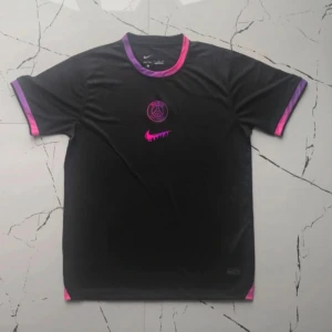 Svart PSG fotbollströja Nike - Snygg svart PSG fotbollströja från Nike med korta ärmar. Tröjan har rosa och lila detaljer vid krage och ärmslut samt PSG-logga och Nike swoosh i neonrosa framtill. Tillverkad i lätt och ventilerande material, perfekt för träning eller match.
