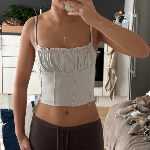 Vit croppad korsett-topp med smala band - Säljer en vit croppad korsett-topp med smala axelband och rynkad överdel. Toppen har synliga sömmar framtill och är figurnära med en snygg, modern look. Perfekt för dig som gillar minimalistisk stil och vill ha något som sticker ut.