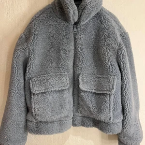 Gråblå teddyjacka med fickor - Mysig grå teddyjacka med fluffigt material och två stora fickor framtill. 