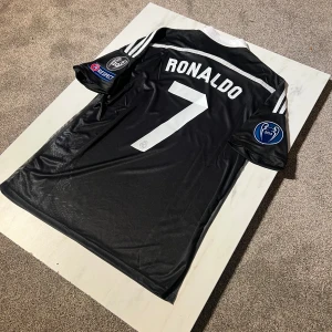 Real Madrid Ronaldo svart CL-tröja - Säljer en svart Real Madrid Champions League-tröja från Adidas med Ronaldo 7 på ryggen. Tröjan har vita detaljer, klubbmärke, Fly Emirates-tryck, CL-patchar och ett drakmönster framtill. Tillverkad i lätt och ventilerande climacool-material.