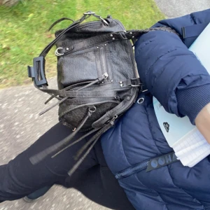 Svart axelväska med dragkedjor - Cool svart axelväska i skinnimitation med flera dragkedjor och långa remmar. Väskan har flätat handtag och snygga metalldetaljer som ger en edgy vibe. Perfekt storlek för att få plats med allt du behöver till skolan eller stan. Har använts ungefär 10 ggr men sen köpte ja en ny väska som ja gillade mer