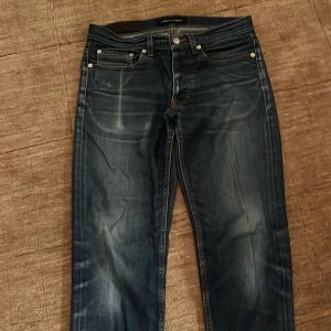 Blå jeans från Sam Mason Denim No.1 - Snygga mörkblå jeans från Sam Mason Denim No.1 med klassisk femficksdesign och kontrastsömmar. Jeansen har raka ben och coola slitningar framtill för en avslappnad vibe. Baktill finns två stora fickor med unik form. Perfekt för dig som gillar stilrena jeans.