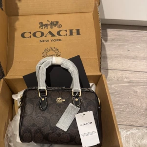 Brun handväska  - Snygg handväska brunt och svart med det klassiska logomönstret. Väskan har guldfärgade detaljer, dubbla handtag och dragkedja upptill. Tillverkad i skinn med stilren och kompakt form, perfekt för dig som gillar lyxiga accessoarer.