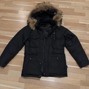 Svart parkajacka med päls från Hollies S - Svart parkajacka från Hollies med snygg fuskpäls på huvan och flera praktiska fickor. Jackan har dragkedja och knappar framtill, samt detaljer i metall. Perfekt för kalla dagar med sin fodrade insida och klassiska look. Storlek 44/S