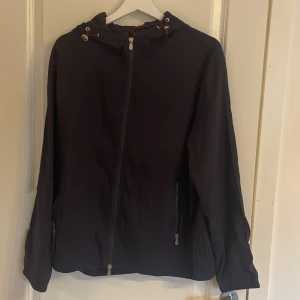 Mörkblå vindjacka med huva - Snygg mörkblå vindjacka med huva och snörning. Jackan har dragkedja framtill och två sidofickor med dragkedja. Passformen är normal och jackan är långärmad, perfekt för dig som gillar enkel och clean stil. Han på bilden är 177