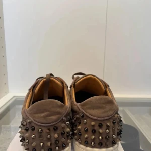 Bruna Louboutin sneakers med nitar storlek 45 - Säljer ett par bruna sneakers från Christian Louboutin i mocka med coola svarta och silvriga nitar på tå och häl. Skorna har snörning och vit platt sula, samt rund tå. Insidan är beige och märkt med Louboutin-loggan. Perfekt för dig som vill sticka ut.