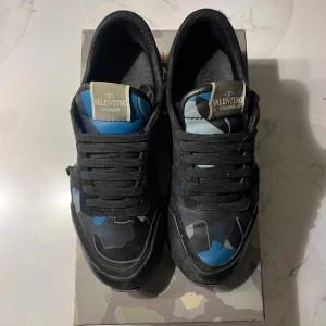 Valentino rockrunner storlek 41, passar 42  - Snygga Valentino Garavani Rockrunner sneakers med svart och blått kamouflagemönster. Skorna har svarta snören, platt sula och detaljer i både skinn och mocka. Ikoniska nitar på hälen och coola kontrasterande paneler gör dem riktigt unika.