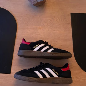 Adidas Handball Spezial svarta sneakers - Adidas Handball Spezial sneakers i svart mocka med vita ränder och bruna gummisulor. Skorna har röda detaljer vid hälen och guldfärgad Spezial-logga på sidan. Klassisk rund tå och snörning framtill. Perfekt för dig som gillar retrostil och streetwear.