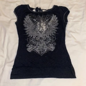 Svart topp med silvertryck och knyt - Cool svart topp med silvrigt tryck. Toppen har korta puffärmar, v-ringad rygg med snygg knyt. Materialet känns mjukt och stretchigt, perfekt för dig som gillar edgy stil med lite extra detaljer!💕