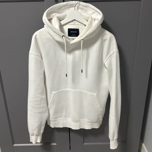 Vit hoodie från Jack & Jones - Väldigt enkel vit hoodie från Jack & Jones med klassisk känguruficka och dragsko i huvan. Tröjan har en relaxed passform och är tillverkad i mjuk bomull, perfekt för chill dagar. Enkel och stilren design som funkar till det mesta.