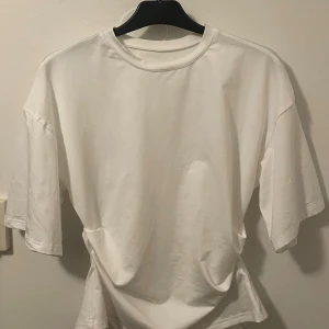 Vit t-shirt från Musera, storlek M - Vit t-shirt från Musera i storlek M. Modellen har en klassisk rund hals och korta ärmar. Materialet är mjukt och lätt, tillverkat av 90% polyester och 10% elastan. T-shirten har en enkel och clean look med en liten slits baktill för extra stil.