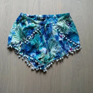 Blommiga shorts med snörning från Bik Bok - Säljer ett par färgglada blommiga shorts från Bik Bok i storlek XS. De har snörning i midjan och vita bollar längs kanten för extra vibe. Mönstret är tropiskt med blå, gröna och vita toner. Perfekta för sommaren och chill dagar.