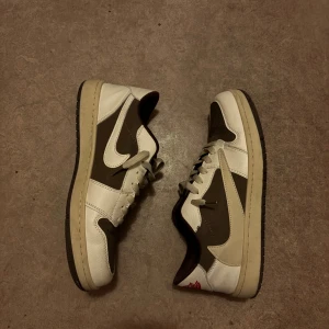 Nike Air Jordan 1 Low Travis Scott Mocha Reverse - Säljer ett par Nike Air Jordan 1 Low Travis Scott med ikonisk omvänd swoosh. Skorna är i brunt och vitt skinn med perforerad tå och klassisk snörning. Röd Nike Air-logga på plösen och broderad detalj på hälen. Perfekt för dig som gillar streetwear och sneakers med hype.