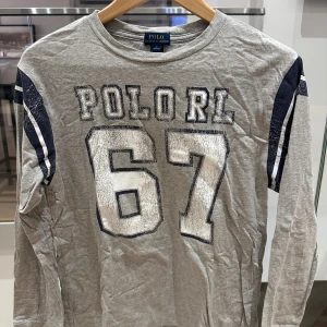 Grå långärmad tröja Polo Ralph Lauren storlek 14-16 år  - Snygg grå långärmad tröja från Polo Ralph Lauren med stort tryck 'POLORL 67' på bröstet och mörkblå ränder på ärmarna. Perfekt för dig som gillar sportig och avslappnad stil. Mjuk bomullskvalitet och rund hals.