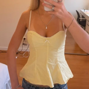 Gul korsett-topp med rosett - Säljer en ljusgul korsett-topp med axelband och söt liten rosett framtill. Toppen har en figurnära passform med markerad midja och lätt utsvängd nederdel. Materialet känns som bomull och toppen har en feminin, fräsch vibe som passar perfekt till jeans.