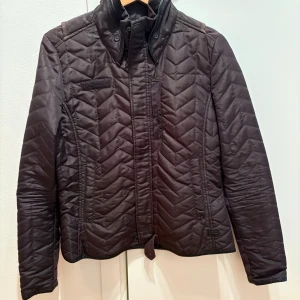 Mörkgrön quiltad jacka från G-Star RAW - Quiltad svart jacka från G-Star RAW Women Raw Cargo Line i storlek M. Jackan har ett snyggt mönster med chevron-sömmar, hög krage och knappar framtill. Materialet är polyester och jackan har långärmade ärmar. Perfekt för höst och vår. Jackan är sparsamt använd, men det finns slitage i ”läderdetaljerna” i nacke och ärmslut pga torka. 