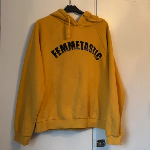 Gul hoodie FEMMETASTIC Gina Tricot - Gul hoodie från Gina Tricot med svart tryck 'FEMMETASTIC' över bröstet. Tröjan har huva med snörning och är tillverkad i mjuk bomullsmix. Perfekt för dig som vill sticka ut med en färgglad och bekväm stil.