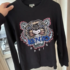 Svart Kenzo tröja med tigerbrodyr - Svart tröja från Kenzo med ikonisk tigerbrodyr framtill i flera färger och texten 'Kenzo Paris'. Tröjan har rund halsringning och långa ärmar. 