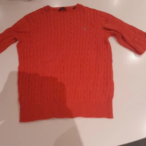 Röd kabelstickad tröja Gant - Snygg röd kabelstickad tröja från Gant med rund halsringning och ribbade muddar. Klassisk design med diskret logga på bröstet. Perfekt för dig som gillar en stilren och färgstark look.