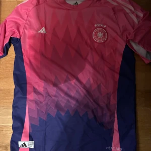 Tysklands fotbollströja rosa/blå Adidas - Säljer en officiell Tyskland fotbollströja från Adidas i rosa och mörkblått med grafiskt mönster. Tröjan har korta ärmar, rund hals och broderat landslagsmärke samt Adidas-logga. Materialet är lätt och andas, perfekt för träning eller match.